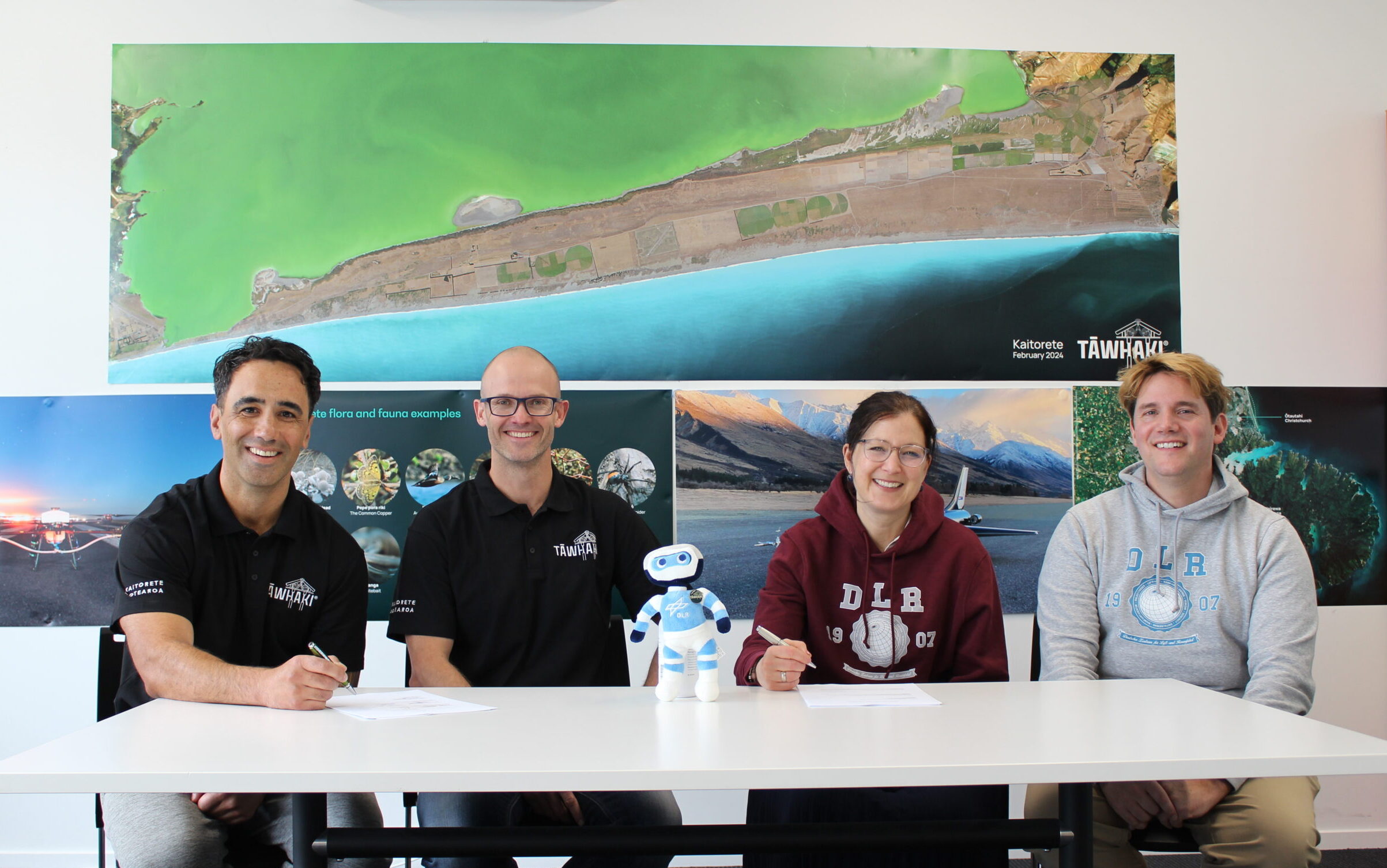 Four people sit at a table to sign an agreement. Two men on the left wearing Tāwhaki shirts, and a woman and a man on the right wearing DLR sweatshirts
